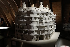 A detailed architectural model of a building with a wavy facade, multiple balconies, and elaborate decorative elements sits under a softly lit environment. The model is monochromatic, primarily white, and features intricate designs that highlight its artistic and architectural features. The background consists of curved, exposed brickwork adding texture and depth.
