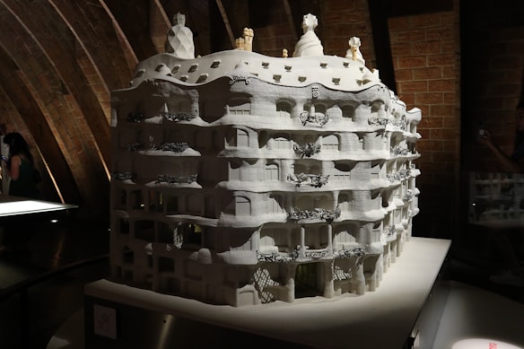 A detailed architectural model of a building with a wavy facade, multiple balconies, and elaborate decorative elements sits under a softly lit environment. The model is monochromatic, primarily white, and features intricate designs that highlight its artistic and architectural features. The background consists of curved, exposed brickwork adding texture and depth.