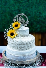 A two-tiered white wedding cake with delicate swirled frosting is adorned with bright yellow sunflowers and small white flowers. A 'Mr & Mrs' cake topper adds a romantic touch, while the cake sits on a rustic wooden stand. The background features a green textured wall, and there are accents of red and white flowers at the base.