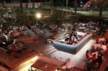 An outdoor, well-lit dining area with people sitting and socializing at various tables. The space features wooden decking and a central bar surrounded by festive lights and foliage. Patrons appear to be enjoying food and drinks under a canopy of trees, with more seating arrangements visible in the background.