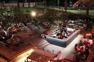 An outdoor, well-lit dining area with people sitting and socializing at various tables. The space features wooden decking and a central bar surrounded by festive lights and foliage. Patrons appear to be enjoying food and drinks under a canopy of trees, with more seating arrangements visible in the background.