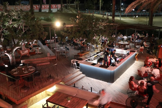 An outdoor, well-lit dining area with people sitting and socializing at various tables. The space features wooden decking and a central bar surrounded by festive lights and foliage. Patrons appear to be enjoying food and drinks under a canopy of trees, with more seating arrangements visible in the background.