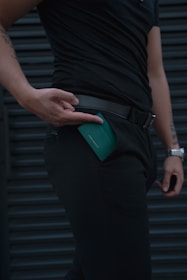 A person dressed in black clothing is holding a green wallet, which is partially inserted into a pocket. The focus is on the hand, wallet, and clothing, creating a sleek and minimalist look. The background features a dark, horizontal pattern.