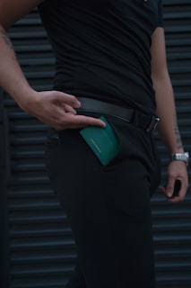 A person dressed in black clothing is holding a green wallet, which is partially inserted into a pocket. The focus is on the hand, wallet, and clothing, creating a sleek and minimalist look. The background features a dark, horizontal pattern.