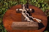 A wooden cross lies on a round, wooden tabletop surrounded by green leaves. The cross features thick brown rope intertwined around it with black fasteners. The base of the cross is inscribed with the phrase 'A cord of three strands is not easily broken. ECCLESIASTES 4:9-12.' The sunlight creates a warm and serene atmosphere.
