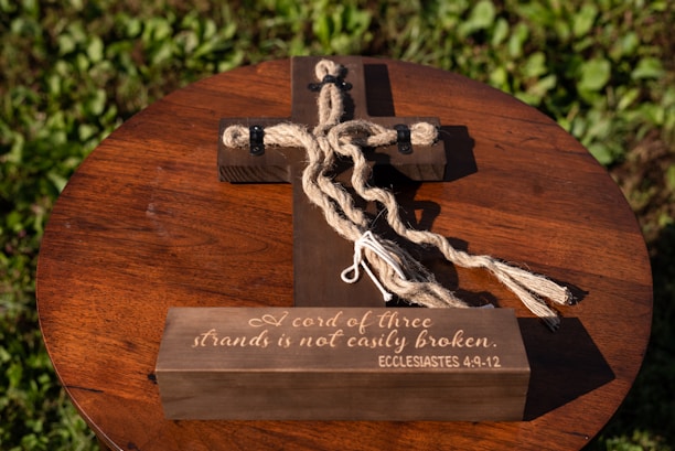 A wooden cross lies on a round, wooden tabletop surrounded by green leaves. The cross features thick brown rope intertwined around it with black fasteners. The base of the cross is inscribed with the phrase 'A cord of three strands is not easily broken. ECCLESIASTES 4:9-12.' The sunlight creates a warm and serene atmosphere.