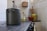A vintage kitchen scene with a large metal pot on a stove. To the right, a collection of kitchenware includes a yellow glass bottle, a red clay pot, and wooden utensils in a decorative yellow-green ceramic jug. The setting includes tiled walls and a small wooden shelf.