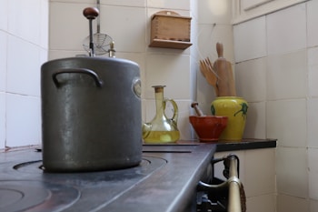 A vintage kitchen scene with a large metal pot on a stove. To the right, a collection of kitchenware includes a yellow glass bottle, a red clay pot, and wooden utensils in a decorative yellow-green ceramic jug. The setting includes tiled walls and a small wooden shelf.