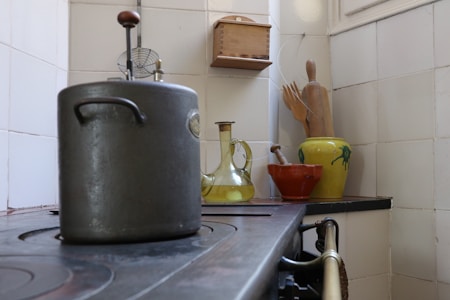 A vintage kitchen scene with a large metal pot on a stove. To the right, a collection of kitchenware includes a yellow glass bottle, a red clay pot, and wooden utensils in a decorative yellow-green ceramic jug. The setting includes tiled walls and a small wooden shelf.