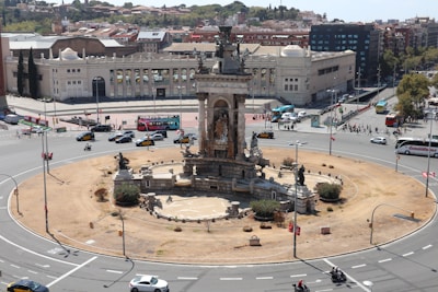 A large, ornate monument stands in the center of a busy roundabout, surrounded by cars, buses, and motorcycles. The monument features intricate sculptures and is set in a circular, landscaped area with low vegetation and pathways. Surrounding buildings include a grand structure with columns that has a historical or government appearance. People are visible on the sidewalks, and the sky is clear, indicating a sunny day.