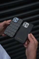 A person holds two smartphones, each in a sleek black case, against a dark, ribbed background. The cases have a luxurious texture, with one featuring a crocodile pattern and the other a smooth finish, both embossed with the brand name. The triple-camera setups are prominently displayed.