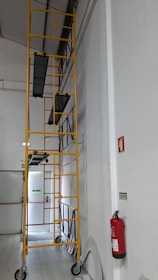 A tall yellow scaffolding structure with multiple platforms is positioned against a white interior wall. The scaffolding is on wheels for mobility within the space. Nearby, a red fire extinguisher is mounted on the wall, and emergency exit signs are visible in the background.