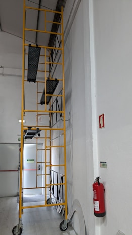 A tall yellow scaffolding structure with multiple platforms is positioned against a white interior wall. The scaffolding is on wheels for mobility within the space. Nearby, a red fire extinguisher is mounted on the wall, and emergency exit signs are visible in the background.