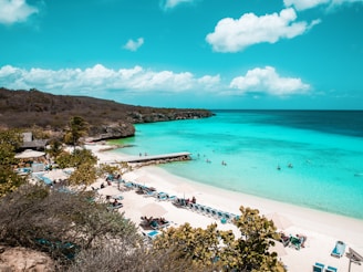 A scenic beach with turquoise waters, white sandy shores, and sun loungers under umbrellas. People are swimming and sunbathing. Green foliage and rocky hills frame the beach, with a clear blue sky overhead.