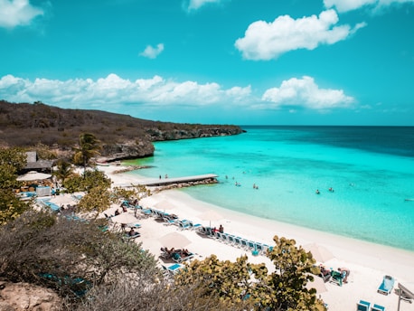 A scenic beach with turquoise waters, white sandy shores, and sun loungers under umbrellas. People are swimming and sunbathing. Green foliage and rocky hills frame the beach, with a clear blue sky overhead.