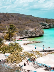 A picturesque beach scene features clear turquoise waters meeting a sandy shore. Sun loungers and umbrellas are scattered along the beach, with several people relaxing or walking around. A palm tree and other greenery add to the tropical aesthetic, while rocky cliffs frame the scene in the background.