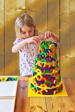 A child is intently building a colorful, pyramid-like tower using interlocking pieces on a wooden table. The background features a rustic wooden wall, and the child is wearing a white shirt with red patterns.