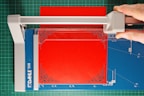 A paper cutter with a blue cutting mat and a red sheet of paper. A hand is visible, using the cutter to trim the paper, which has ornate corner designs. The cutting surface has measurement markings, and the cutter blade is housed in a grey plastic mechanism.