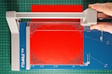 A paper cutter with a blue cutting mat and a red sheet of paper. A hand is visible, using the cutter to trim the paper, which has ornate corner designs. The cutting surface has measurement markings, and the cutter blade is housed in a grey plastic mechanism.
