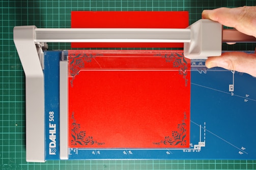 A paper cutter with a blue cutting mat and a red sheet of paper. A hand is visible, using the cutter to trim the paper, which has ornate corner designs. The cutting surface has measurement markings, and the cutter blade is housed in a grey plastic mechanism.
