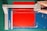 A paper cutter with a blue cutting mat and a red sheet of paper. A hand is visible, using the cutter to trim the paper, which has ornate corner designs. The cutting surface has measurement markings, and the cutter blade is housed in a grey plastic mechanism.