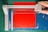 A paper cutter with a blue cutting mat and a red sheet of paper. A hand is visible, using the cutter to trim the paper, which has ornate corner designs. The cutting surface has measurement markings, and the cutter blade is housed in a grey plastic mechanism.