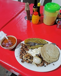 A plate of Mexican food on a red table. The dish includes grilled onions, beef, a serving of cheese, corn tortillas, and a cactus paddle. A bowl of red salsa with onions is placed nearby, and various condiments including sauces and napkins are on the table.