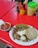 A plate of Mexican food on a red table. The dish includes grilled onions, beef, a serving of cheese, corn tortillas, and a cactus paddle. A bowl of red salsa with onions is placed nearby, and various condiments including sauces and napkins are on the table.