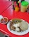 A plate of Mexican food on a red table. The dish includes grilled onions, beef, a serving of cheese, corn tortillas, and a cactus paddle. A bowl of red salsa with onions is placed nearby, and various condiments including sauces and napkins are on the table.