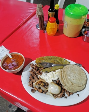 A plate of Mexican food on a red table. The dish includes grilled onions, beef, a serving of cheese, corn tortillas, and a cactus paddle. A bowl of red salsa with onions is placed nearby, and various condiments including sauces and napkins are on the table.
