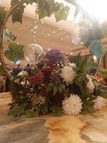 A decorative arrangement featuring vibrant flowers and lush green leaves on a table. There is a wooden arch adorned with hanging glass globes and vines, creating an elegant display. The background shows a softly lit room.