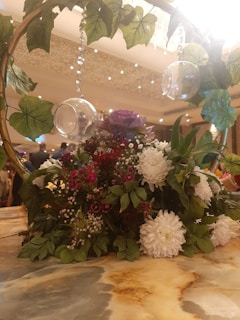 A decorative arrangement featuring vibrant flowers and lush green leaves on a table. There is a wooden arch adorned with hanging glass globes and vines, creating an elegant display. The background shows a softly lit room.