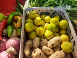 A variety of fresh vegetables and fruits are arranged in a market setting. There are bright yellow lemons in a white plastic basket, surrounded by root vegetables, green peppers, red peppers, and purple onions. In the background, there are cucumbers and green beans. The scene is well-lit, highlighting the vibrant colors of the produce.