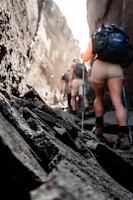 A group of hikers with backpacks navigates a narrow rocky passage between steep cliffs. The sunlight filters through the crevices, casting a warm glow on the scene. The rugged terrain is made of dark stones and rocks, with the hikers wearing casual outdoor attire.