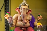 A colorful statue of the Hindu deity Ganesha, depicted with a large elephant head and four arms. The statue is adorned with intricate jewelry and wears a purple drape over one shoulder. One hand holds a yellow sweet, while the others hold red and pink flowers. The figure sits in a relaxed pose on a shelf, surrounded by a soft yellow background.