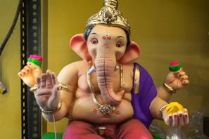 A colorful statue of the Hindu deity Ganesha, depicted with a large elephant head and four arms. The statue is adorned with intricate jewelry and wears a purple drape over one shoulder. One hand holds a yellow sweet, while the others hold red and pink flowers. The figure sits in a relaxed pose on a shelf, surrounded by a soft yellow background.
