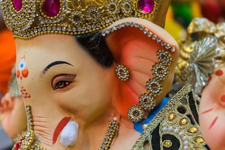 A detailed and colorful sculpture adorned with intricate beadwork and jewels, featuring an elephant-headed figure with a serene expression. The decoration includes vibrant colors, with gold accents and patterns, showcasing traditional artistic craftsmanship.