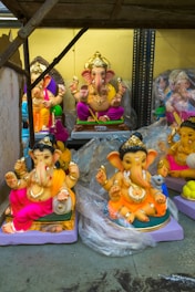 A collection of colorful statues of the Hindu deity Ganesha, placed on shelves. Each statue depicts Ganesha in a seated position, adorned with gold jewelry, vibrant clothing, and ornate crowns. Some statues are wrapped in clear protective plastic.
