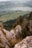 A scenic landscape with a high vantage point showcasing a vast valley below, filled with fields and a serene lake bordered by mountains. Rocky cliffs and dense green forests are prominent in the foreground, with a lone person climbing the rocks, adding a sense of adventure.