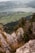 A scenic landscape with a high vantage point showcasing a vast valley below, filled with fields and a serene lake bordered by mountains. Rocky cliffs and dense green forests are prominent in the foreground, with a lone person climbing the rocks, adding a sense of adventure.