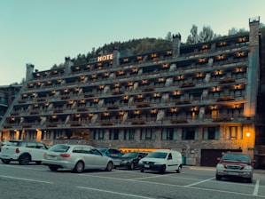 A multi-story hotel building with numerous balconies is situated in front of a parking lot filled with various cars. The structure has a rustic appearance with stone and brickwork, and warm lighting illuminates the exterior. Behind the hotel, a forested hillside rises, contributing to a serene setting.
