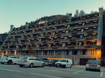 A multi-story hotel building with numerous balconies is situated in front of a parking lot filled with various cars. The structure has a rustic appearance with stone and brickwork, and warm lighting illuminates the exterior. Behind the hotel, a forested hillside rises, contributing to a serene setting.