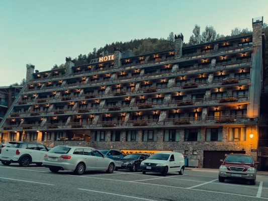 A multi-story hotel building with numerous balconies is situated in front of a parking lot filled with various cars. The structure has a rustic appearance with stone and brickwork, and warm lighting illuminates the exterior. Behind the hotel, a forested hillside rises, contributing to a serene setting.