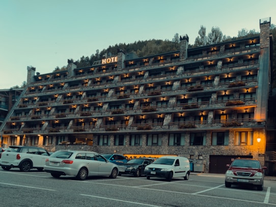 A multi-story hotel building with numerous balconies is situated in front of a parking lot filled with various cars. The structure has a rustic appearance with stone and brickwork, and warm lighting illuminates the exterior. Behind the hotel, a forested hillside rises, contributing to a serene setting.