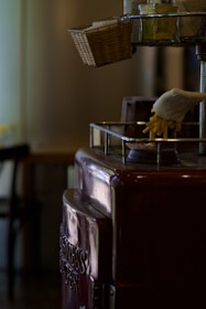 A vintage metal popcorn machine with a glossy maroon finish is situated indoors. An artificial hand is poised over the machine with artificial popcorn spilling from its grip. A wicker basket is attached to the top corner, and there are containers of seasoning or condiments next to it. The background appears to be a dimly lit room.