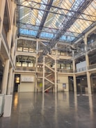 A large, open interior space with multiple floors featuring a modern architectural design. The space includes industrial elements like metal beams and a glass ceiling that allows natural light to flood in. There is a prominent central staircase and balconies on different levels, with signs and posters visible on the walls.