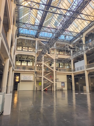 A large, open interior space with multiple floors featuring a modern architectural design. The space includes industrial elements like metal beams and a glass ceiling that allows natural light to flood in. There is a prominent central staircase and balconies on different levels, with signs and posters visible on the walls.