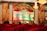 A stage setup for a dance competition featuring a large backdrop with ornate golden designs and a banner displaying the words 'Dance Competition' in bold letters. The stage is decorated with red drapery and floral arrangements. Chandeliers hang from the ceiling, and photography or video equipment is set up around the stage.