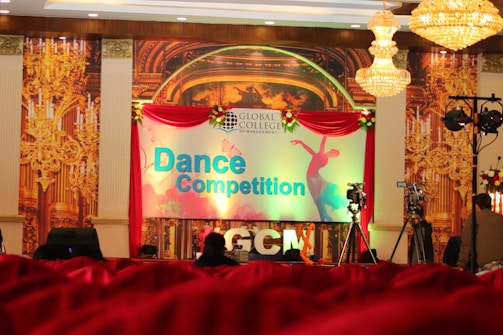 A stage setup for a dance competition featuring a large backdrop with ornate golden designs and a banner displaying the words 'Dance Competition' in bold letters. The stage is decorated with red drapery and floral arrangements. Chandeliers hang from the ceiling, and photography or video equipment is set up around the stage.