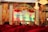 A stage setup for a dance competition featuring a large backdrop with ornate golden designs and a banner displaying the words 'Dance Competition' in bold letters. The stage is decorated with red drapery and floral arrangements. Chandeliers hang from the ceiling, and photography or video equipment is set up around the stage.